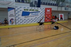 Zdjęcie przedstawia żółtą łódź wioślarską eksponowaną w hali sportowej na tle banerów promujących sprzęt wioślarski oraz fundację Row Sport Fan.