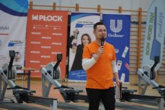 Zdjęcie przedstawia prowadzącego w pomarańczowej koszulce mówiącego do mikrofonu podczas wydarzenia sportowego przy ergometrach wioślarskich.