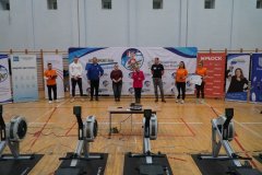 Grupa dorosłych stoi na sali gimnastycznej przed banerami i ergometrami.