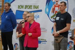 Dyrektor Szkoły Pani Marlena Błachowicz w różowej marynarce przemawia do mikrofonu, stojąc z grupą osób przed banerem wydarzenia sportowego.