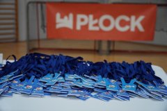 Na stole leżą liczne niebieskie medale, a w tle widnieje baner z napisem „Płock”.