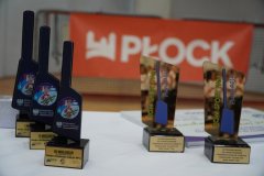 Na stole stoją trofea o tematyce sportowej, a w tle widać baner z napisem „Płock”.