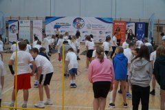 Dzieci w szkolnej sali gimnastycznej wykonują ćwiczenia pod kierunkiem instruktorów przy banerach sportowego wydarzenia.