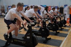 Grupa dzieci ćwiczy na rowerach stacjonarnych w sali sportowej podczas szkolnego wydarzenia.