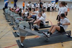 Dzieci ćwiczą na ergometrach wioślarskich w dużej sali gimnastycznej podczas sportowych zajęć.