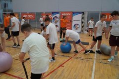Grupa uczniów ćwiczy z piłkami gimnastycznymi na sali sportowej pod okiem instruktorów.