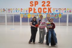 W przestronnej sali z dużym, pomarańczowym napisem "SP 22 Płock" oraz kolorowymi postaciami dzieci oraz dużymi wielobarwnymi pisankami z kartonu w tle cztrey uczennice klas 4-8 ze wsoimi zamiennikami plecaków (pluszowy koszyczek na zakupy, ogromny pluszowy koc-worek, walizka i opakowanie po kapsułkach piorących).