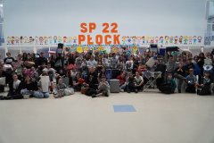 W przestronnej sali z dużym, pomarańczowym napisem "SP 22 Płock" oraz kolorowymi postaciami dzieci oraz dużymi wielobarwnymi pisankami z kartonu w tle znajduje sie bardzo duża grupa uczniów klas 4-8. Wszyscy z bardzo wymyślnymi zamiennikami plecaków (np. suszarka na pracnie, hamak, garnki, pojemniki na ciasto, nocnik, walizki, kosze)