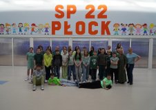 W przestronnej sali z dużym, pomarańczowym napisem "SP 22 Płock" oraz kolorowymi postaciami dzieci w tle znajduje się grupa uczniów klas 4-8 wraz dyrektorką i nauczycielką, wszyscy ubrani na zielono i w pogodnych nastrojach.