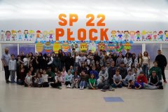 W przestronnej sali z dużym, pomarańczowym napisem "SP 22 Płock" oraz kolorowymi postaciami dzieci oraz dużymi wielobarwnymi pisankami z kartonu w tle znajduje się kilka rzędów uczniów klas 4-8 oraz nauczycieli, wszyscy w ubraniach na opak