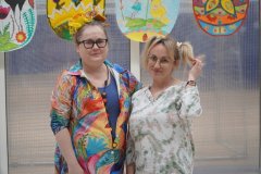 Dwie nauczycielki w szlonych fryzurach, jedna z artystycznymi dodatkami na głowie: pędzelkami o różnych rozmiarach oraz innymi dekoracjami, a druga trzyma się za jedną z dwóch rozpuszonych kitek. W tle fragmenty dużych, kolorowych, ozdobnych pisanek z papieru.