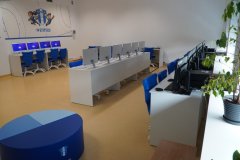 Rząd stanowisk komputerowych z niebieskimi krzesłami i fioletowymi ekranami w jasnej sali lekcyjnej.