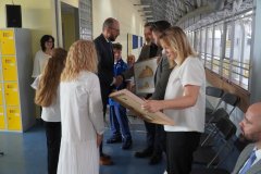 Uczestnicy uroczystości wymieniają uściski dłoni i wręczają pamiątkowe, oprawione obrazy na szkolnym korytarzu w obecności dzieci i zaproszonych gości.