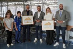 Oficjalne podziękowania w szkole; dorośli goście oraz dwoje dzieci pozują do zdjęcia, trzymając w rękach dwa oprawione w ramy, drewniane obrazy przedstawiające panoramę miasta.