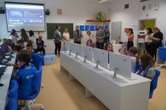 Uczniowie i dorośli zgromadzeni w nowoczesnej sali komputerowej z rzędami białych biurek; na ścianie wyświetlany jest obraz z projektora multimedialnego.