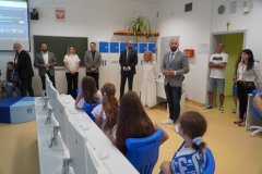 Mężczyzna w szarej marynarce przemawia do dzieci siedzących przy biurkach komputerowych, podczas gdy pozostali zaproszeni goście stoją w rzędzie pod ścianą sali.