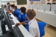 Uczniowie w niebieskich koszulkach sportowych i białych galowych koszulach siedzą przy stanowiskach komputerowych i pracują przy monitorach w nowej pracowni.
