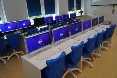 Sala lekcyjna z rzędami komputerów na biurkach, niebieskimi krzesłami, rośliną w doniczce.
