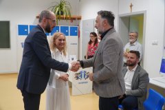 Dwaj mężczyźni w eleganckich strojach wymieniają uścisk dłoni w nowej sali komputerowej, w obecności uśmiechniętej kobiety w białej sukience oraz innych gości.