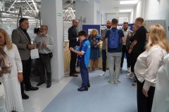 Zgromadzeni goście, dorośli i dzieci, rozmawiają w szkolnym korytarzu podczas spotkania; w tle widoczny jest bufet cateringowy oraz niebieska tablica dekoracyjna.