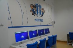 Sala lekcyjna z rzędami komputerów na biurkach, niebieskimi krzesłami, oraz ściennym malunkiem przedstawiającym postacie z kreskówek i napis „ENTELO”.