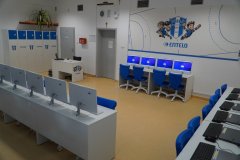 Jasna, nowoczesna pracownia komputerowa z rzędami białych biurek, na których ustawiono monitory z niebieskimi krzesłami obrotowymi.
