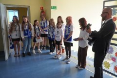 Szkolny chór dziewcząt w kibicowskich szalikach śpiewa w korytarzu, a mężczyzna w okularach i garniturze gra obok nich na gitarze akustycznej.