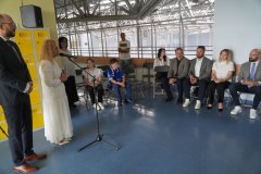 Oficjalne przemówienie kobiety w białej sukience (pani Dyrektor Sp 22) stojącej przy mikrofonie, podczas gdy zgromadzona publiczność i zaproszeni goście słuchają jej, siedząc w rzędzie na krzesłach.