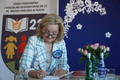 Pani Kurator Wioletta Krzyżanowska podpisuje Konstytucję przy stole, w tle baner Szkoły Podstawowej nr 22 w Płocku, obok kwiaty i woda.