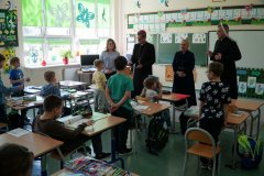 Zdjęcie przedstawia dzieci z klasy 1b znajdujące się w sali lekcyjnej. W głębi od lewej strony widzimy p. katechetkę Edytę Woźniak, z prawej strony ks. Biskupa Mirosława Milewskiego a obok p. dyrektor Marlenę Błachowicz oraz ks. Piotra Szulca.