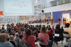 Grupa nauczycieli, uczniów tworzących widownię oraz aktorów ogląda prezentację multimedialną wyświetlaną na jednej ze ścian forum. Jej treść omawia koordynator spotkania- p. Anna Kowalska.