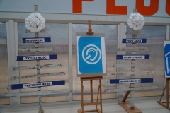 Na tle jednej ze ścian form, na sztalugach, ustawione są piktogramy informujące o różnych rodzajach niepełnosprawności oraz drewniane choinki z hasłami nawołującymi do tolerancji i równości wobec osób z niepełnosprawnościami.