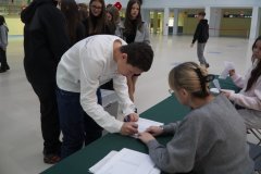 Uczeń podpisuje się na liście wyborców przy stoliku komisji wyborczej podczas wyborów do Młodzieżowej Rady Miasta Płocka w szkolnym holu. W tle widoczni są inni uczniowie oczekujący na oddanie głosu.