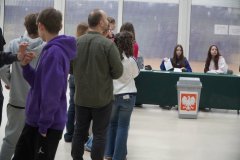 W tle stolik komisji wyborczej podczas wyborów do Młodzieżowej Rady Miasta Płocka w szkolnym holu. Na pierwszym planie są uczniowie oczekujący na oddanie głosu.
