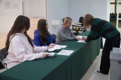 Uczennice przy stoliku przykrytym zieloną tkaniną sprawdzają listę wyborców i wydają karty do głosowania, podczas gdy uczennica podpisuje się na liście w trakcie szkolnych wyborów do Młodzieżowej Rady Miasta Płocka.