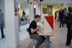 Uczeń oddaje głos w specjalnie przygotowanej kabinie wyborczej ustawionej na terenie szkoły. Wokół znajdują się inni uczniowie oraz elementy przestrzeni szkolnej podczas trwania wyborów.