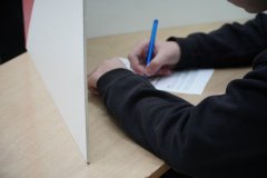 Uczeń oddaje głos w specjalnie przygotowanej biało-czerwonej kabinie wyborczej ustawionej na terenie szkoły. Zbliżenie pokazuje moment wypełniania karty do głosowania w spokojnej, wydzielonej przestrzeni zapewniającej tajność głosowania.