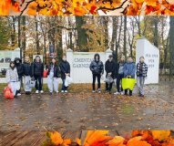 Na zdjęciu widać grupę młodych osób stojących przed wejściem na Cmentarz Garnizonowy. Za nimi znajduje się biały mur z napisem „Cmentarz Garnizonowy” oraz datami „1914–1918, 1920, 1939”. W tle rosną wysokie drzewa w jesiennych barwach. Uczestnicy trzymają reklamówki i torby, prawdopodobnie ze zniczami. Całość otacza dekoracyjna ramka z motywem jesiennych liści oraz drewnianą fakturą u dołu.