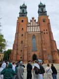 Grupa uczniów stoi przed monumentalną fasadą Katedry Poznańskiej. Budynek z czerwonej cegły, z charakterystycznymi dwiema wieżami. Przewodnik wskazuje na detale architektoniczne.