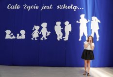 Elegancko ubrana dziewczynka na tle niebieskiej dekoracji z napisem ,,Całe życie jest szkołą” wykonuje utwór muzyczny.