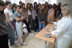 Uczestnicy konkursu zebrani wokół nauczycielki fizyki, która demonstruje doświadczenie o tonięciu i pływaniu ciał, z użyciem miski z wodą i kilku różnych przedmiotów, np. jajka, korka, baterii, patyczka, drucika itp.