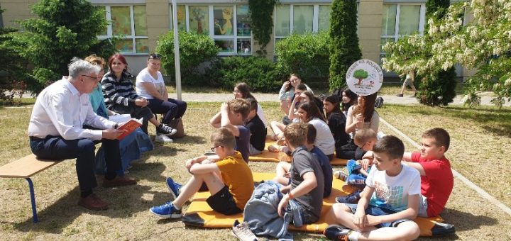 Pan dyrektor czyta na głos grupie uczniów siedzących wokół niego na trawie.