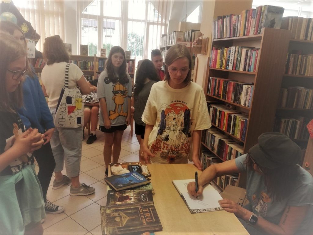 Autorka Barbara Kosmowska pozuje z uczniami klasy 6e do zdjęcia. Wszyscy siedzą w półkolu na krzesłach.