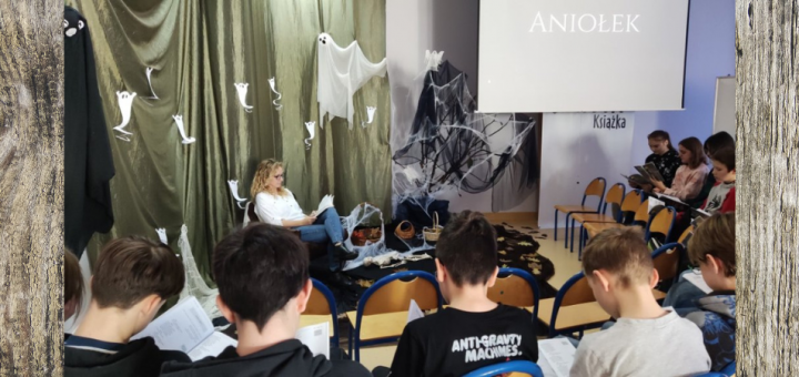 Grupa uczniów czyta II część „Dziadów” Adama Mickiewicza w scenerii biblioteki szkolnej zaaranżowanej specjalnie na tę okoliczność.
