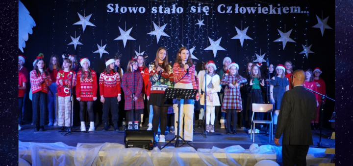 Scena na której stoi chór szkolny, dwie solistki trzymają mikrofony.