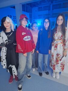 Foto_1 : Na zdjęciu znajduje się grupa pięciu dziewczynek stojących koło siebie, w przebraniach karnawałowych na tle oświetlonego na niebiesko forum Szkoły.