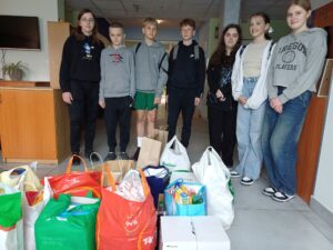 „Grupa siedmiorga młodych osób stoi w pomieszczeniu, uśmiechając się do zdjęcia. Przed nimi znajduje się wiele toreb i reklamówek wypełnionych różnymi produktami, zebranymi w ramach akcji charytatywnej lub zbiórki. W tle widać biuro lub szkolny korytarz z szafkami, rośliną i telewizorem zawieszonym na ścianie.”