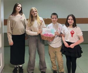 Czworo uczniów ze szkolnego Samorządu Uczniowskiego stoi w korytarzu. Trzymają koszyk wypełniony biało-czerwonymi kotylionami oraz kartkę z napisem „Samorząd Uczniowski”. Na ubraniach mają przypięte kotyliony z okazji Narodowego Święta Niepodległości.