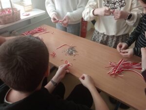 Uczniowie siedzą przy stole i własnoręcznie wykonują biało-czerwone kotyliony z wstążek. Na blacie leżą przygotowane paski wstążek oraz zapinki, a w tle widać koszyk z gotowymi ozdobami. Zdjęcie przedstawia etap przygotowań do obchodów Narodowego Święta Niepodległości.