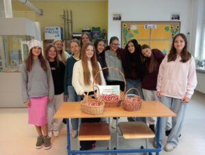 Grupa uczennic ze szkolnego Samorządu Uczniowskiego stoi w klasie za stołem, na którym ustawiono trzy koszyki wypełnione biało-czerwonymi kotylionami oraz kartkę z napisem „Samorząd Uczniowski”. Uczennice uśmiechają się do zdjęcia.